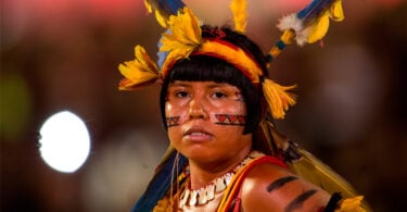 Mulher olhando para frente com traços de tinta no rosto e na cabeça uma tiara com penas ao redor