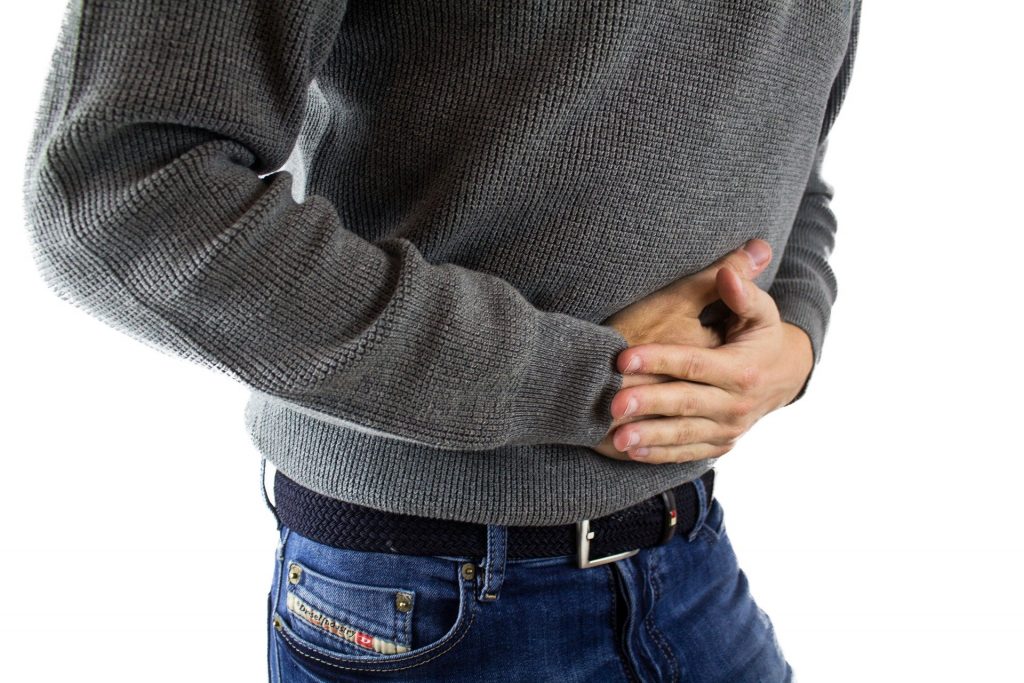 Homem com fortes dores abdominais. Ele está com as duas mãos sobre o abdomen. Ele veste uma blusa de lã cinza e uma calça jeans na cor azul.
