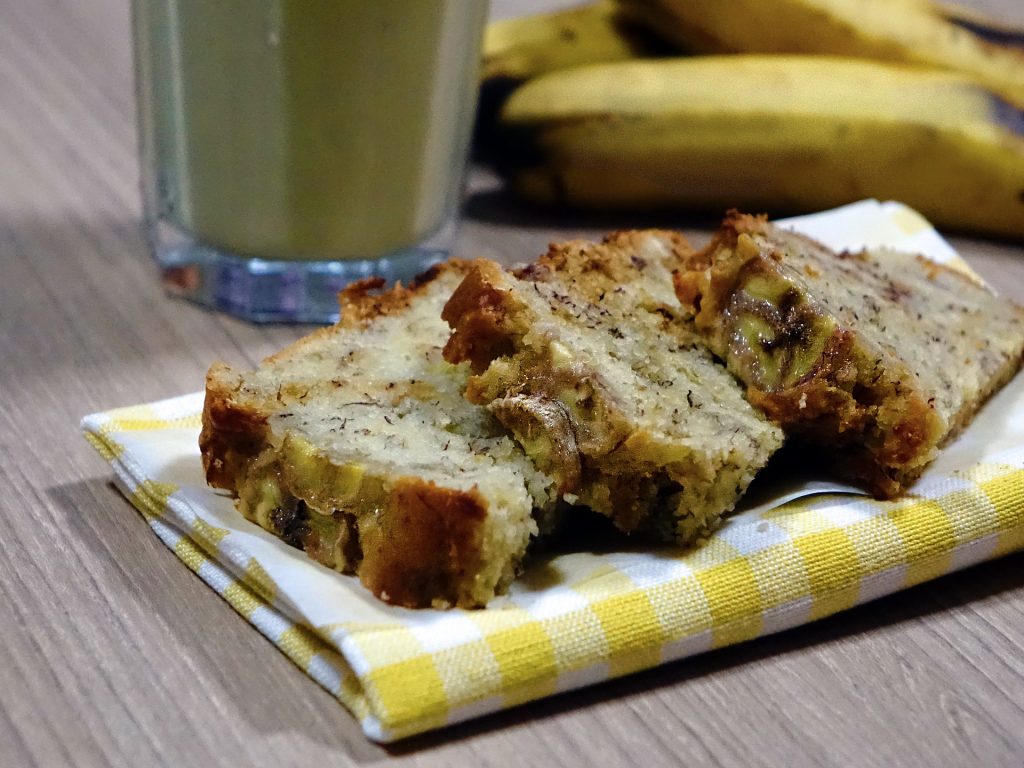 Três fatias de bolo de banana colocadas sobre uma toalha xadrez nas cores amarelo e branco, Ao lado bananas inteira e um copo de suco de banana.