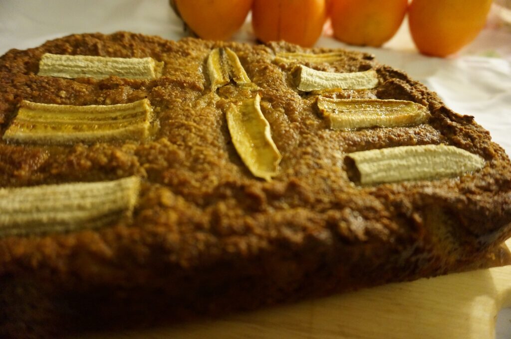 Bolo de banana em formato retangular. Ele já está assado. Sobre ele fatias de bananas assadas.
