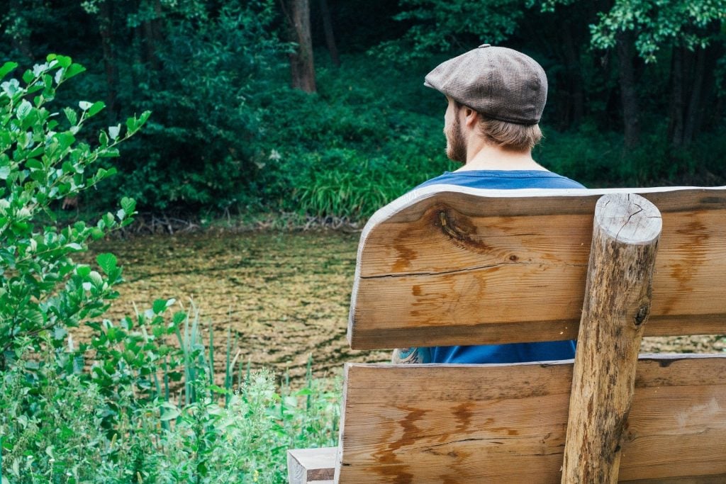 Homem jovem usando barba e boina sentando em um banco de madeira no meio de uma floresta. Ele está em silêncio contemplando a beleza da natureza.
