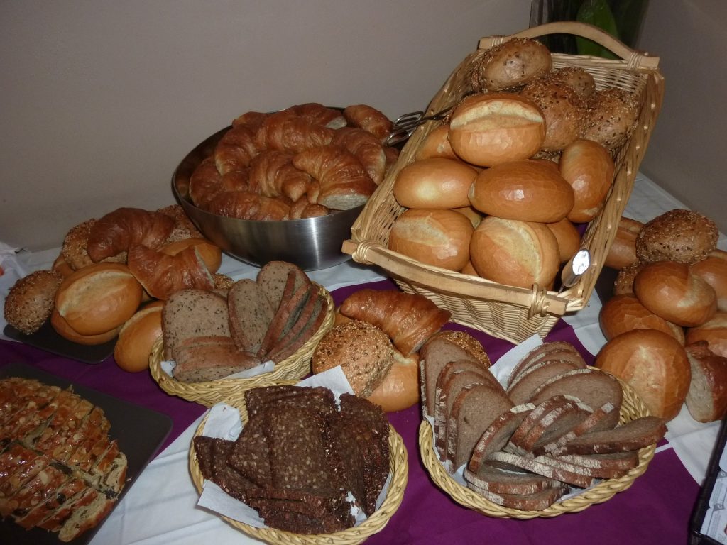 Diversos cestos com variados tipos de pães inntegrais. Os cestos estão distribuídos sobre uma mesa com uma toalha lilás e branca.