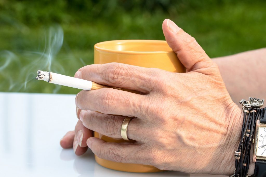 Imagem de mãos de uma mulher segurando uma caneca de café e um cigarro aceso. As mãos estão meio envelhecidas.
