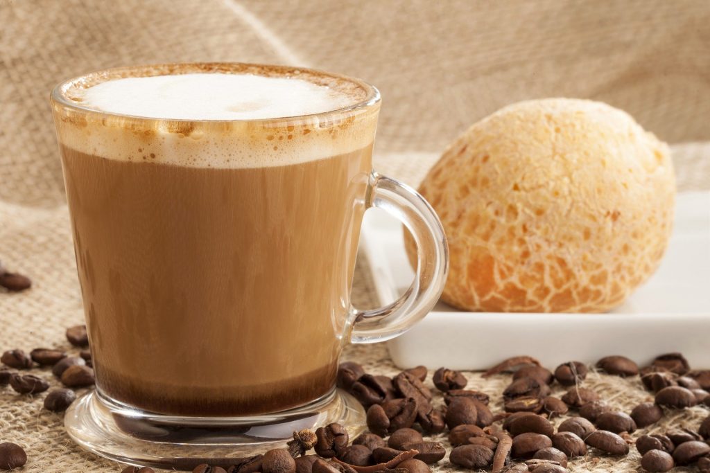 Xícara de vidro com café com leite. Ao lado um pão de queijo dispostos sobre uma mesa decorada com um pano de juta e grãos de café.