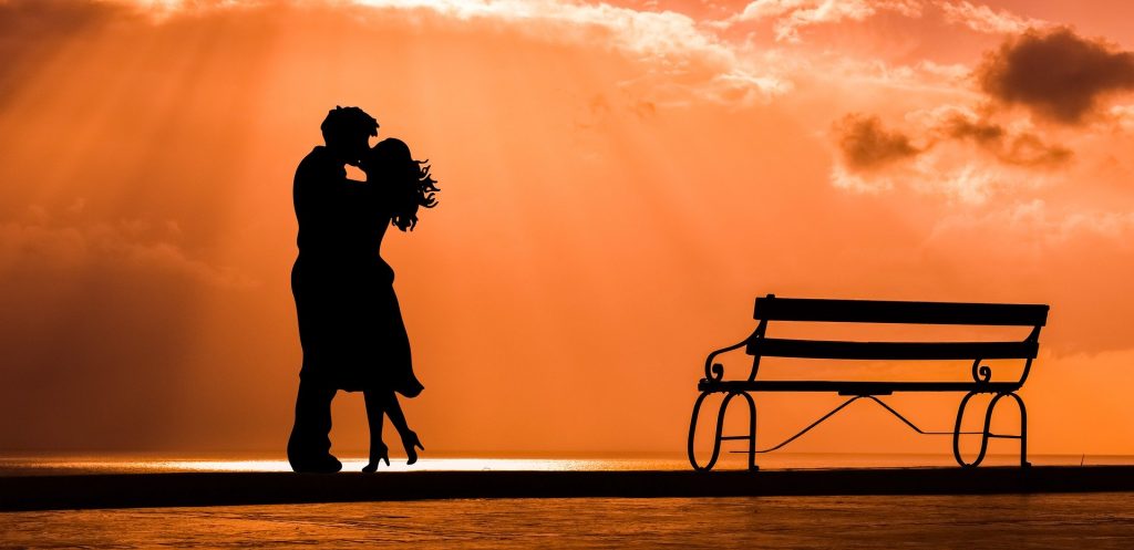 Imagem de um casal homem e mulher. Eles estão se beijando em frente ao mar em um lindo por do sol. Ao lado um banco de ferro compõe a imagem.