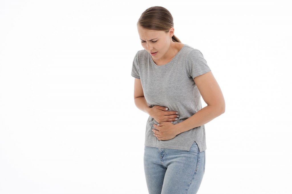 Imagem de uma mulher loura de cabelos longos e amarrados, segurando o estomâgo Ela está cm fortes dores. Ela veste uma camiseta cinza e uma calça jeans na cor clara.