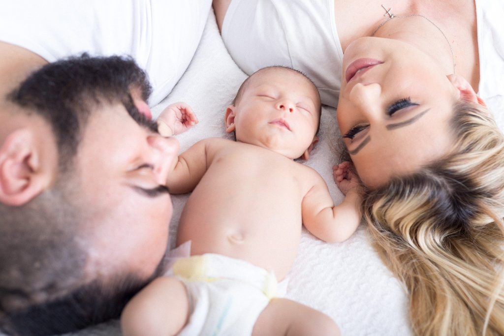 Família composta por um homem e uma mulher e ao centro um bebê recém nascido. Todos estão deitados sobre uma cama e a criança está ao centro.