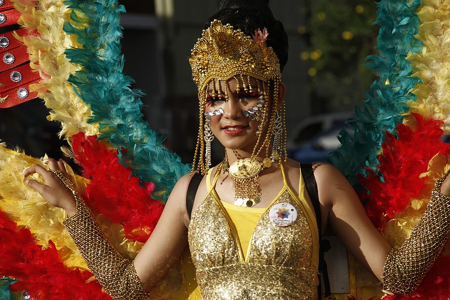 Mulher com fantasia de carnaval dourada, com coroa cheia de pedras douradas e prateadas penduradas, e asas grandes com penas em azul, vermelho e amarelo pendurada em suas costas.