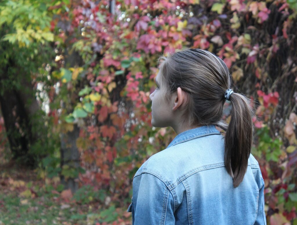 Imagem de uma garota jovem vestindo uma jaqueta jeans azul clara. Ela está em silêncio observando as folhagens que caíram da árvore em uma linda tarde de outono.
