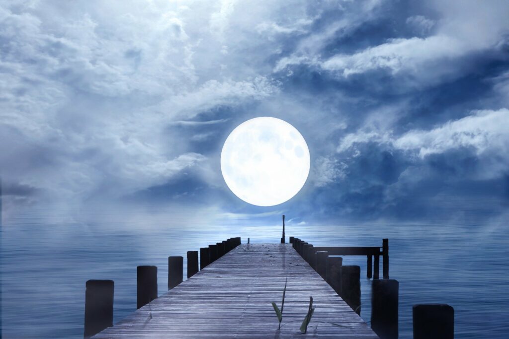 Imagem de uma ponte sobre o mar em um início de noite. Ao fundo a lua chei ilumina a paisagem.