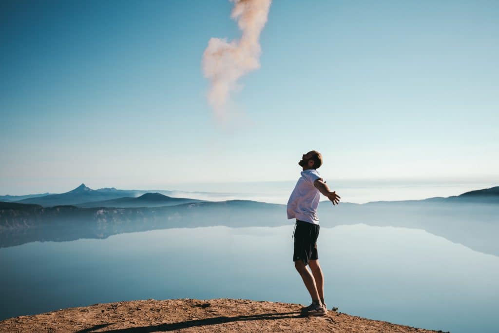 Homem em montanha com braços abertos de lado com mar e céu azul ao fundo