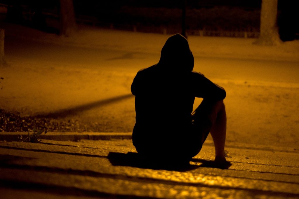Imagem de um  homem sentado na guia de uma calçada. Ele veste uma roupa preta e usa uma touca da blusa sobre a sua cabeça.