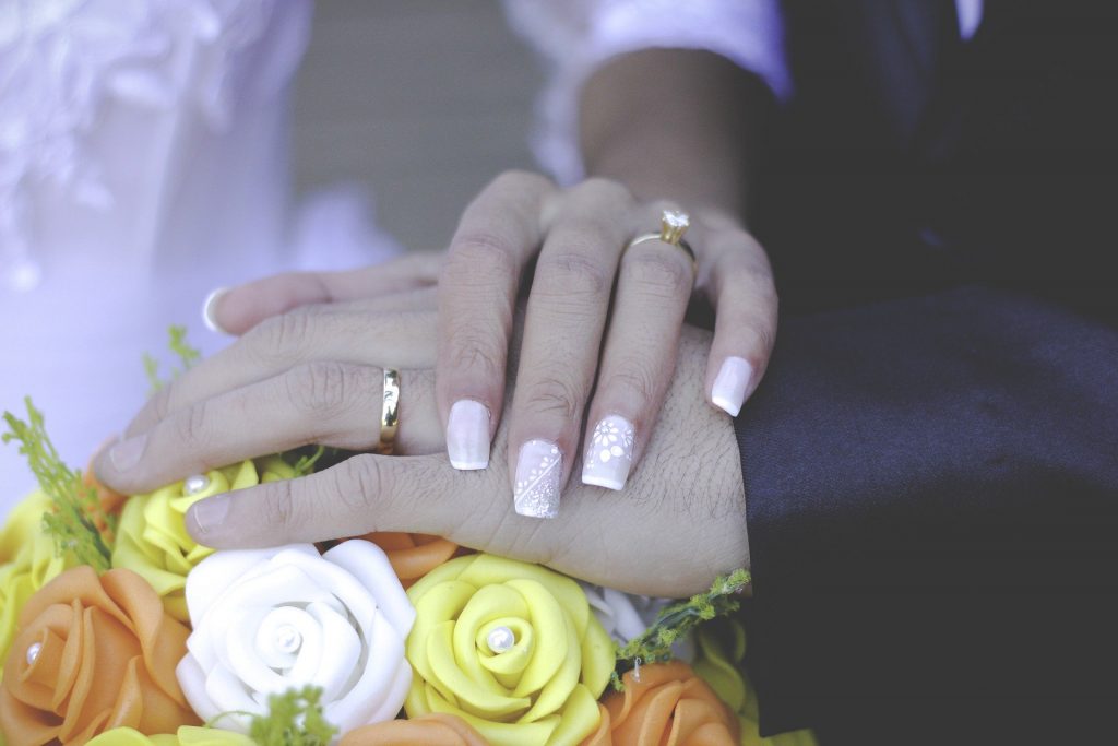 Imagem das mãos de um casal de homem e mulher usando alianças na cerimônia de casamento,
As mãos estão posicionadas sobre o buquê de noiva feito de flores amarelas, brancas e alaranjadas.
