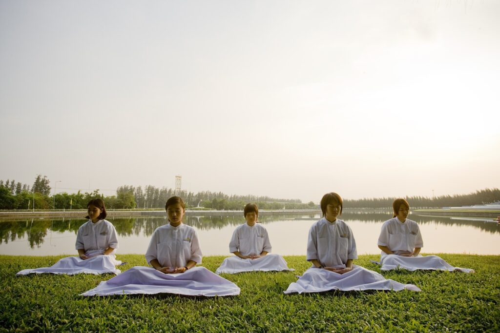 Imagem de um grupo de crianças japonesas meditando em uma escola. São cinco crianças sentadas em um gramado verdinho, todas vestindo roupas brancas.
