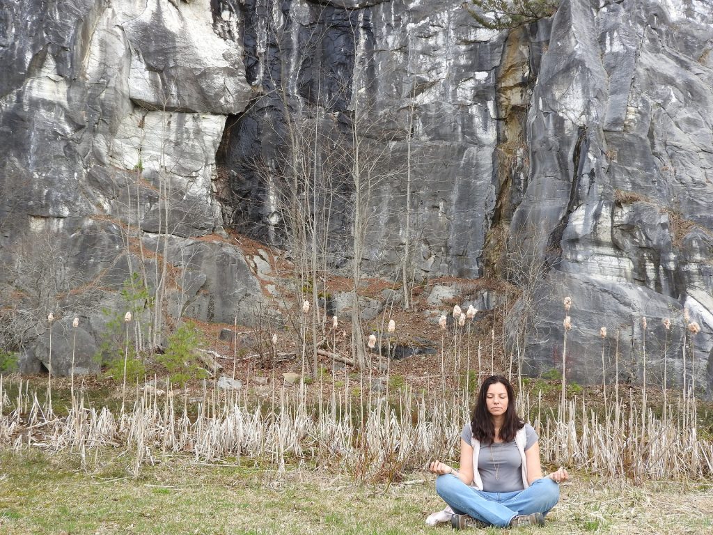Imgem de uma mulher sentada em um gramado em posição de meditação. Ao fundo dela uma parede rochosa. Ela está com os olhos fechados, usa camiseta cinza, colete bege e uma calça jeans.
