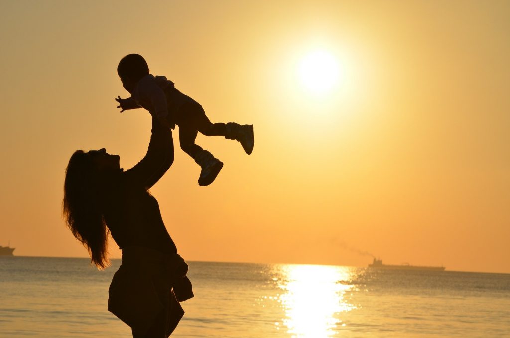 Imagem de uma mãe segurando e levantando o seu filho no colo. Ao fundo a luz brilhante de um lindo por do sol.