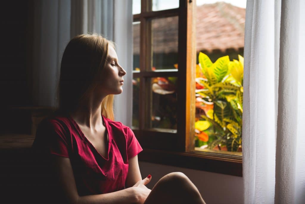 Mulher sentada em frente à janela, de olhos fechados, meditando.