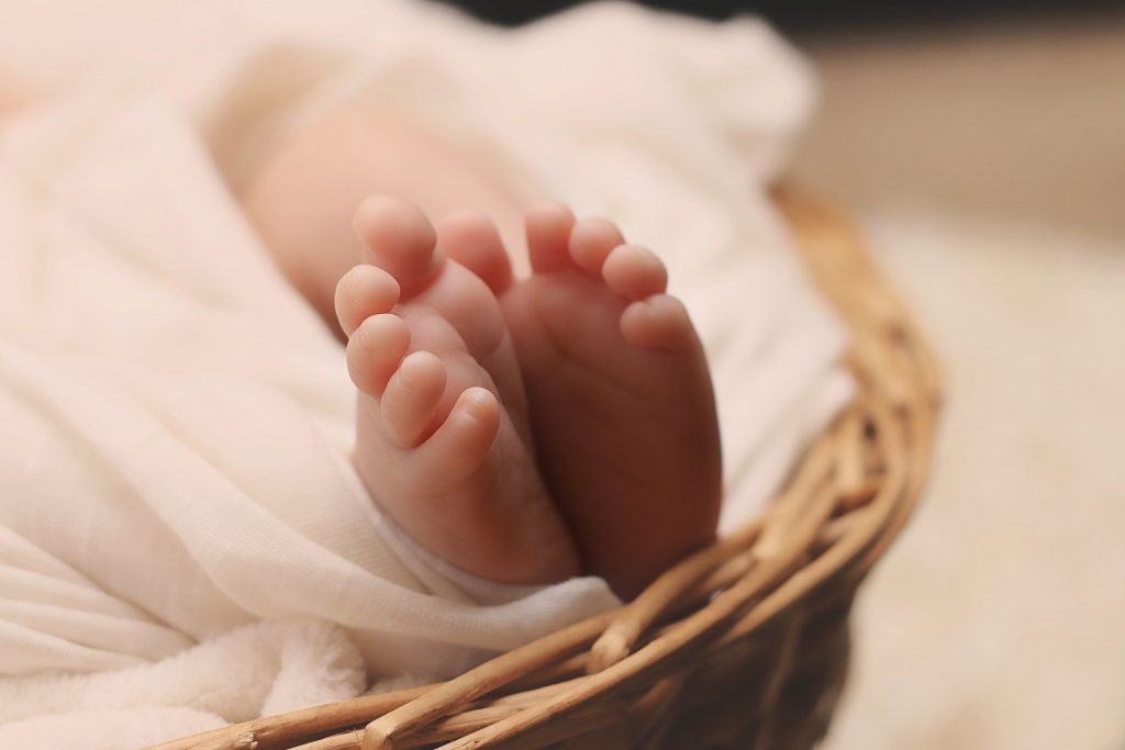 Imagem de um pé de um bebê recém nascido. Ele está deitado dentro de um cesto de vime coberto com um cobertor branco.