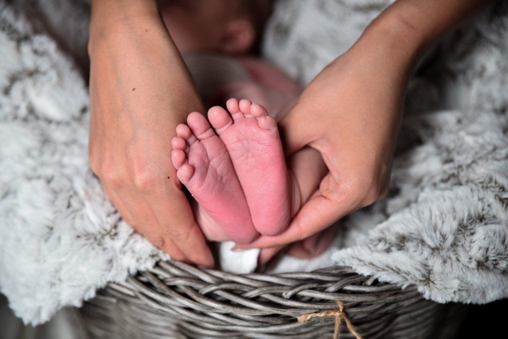 Imagem das mãos da mãe segurando os pés de um bebê recém nascido. O bebê está deitando dentro de um cesto de vime forrado com um manto de pele branco.