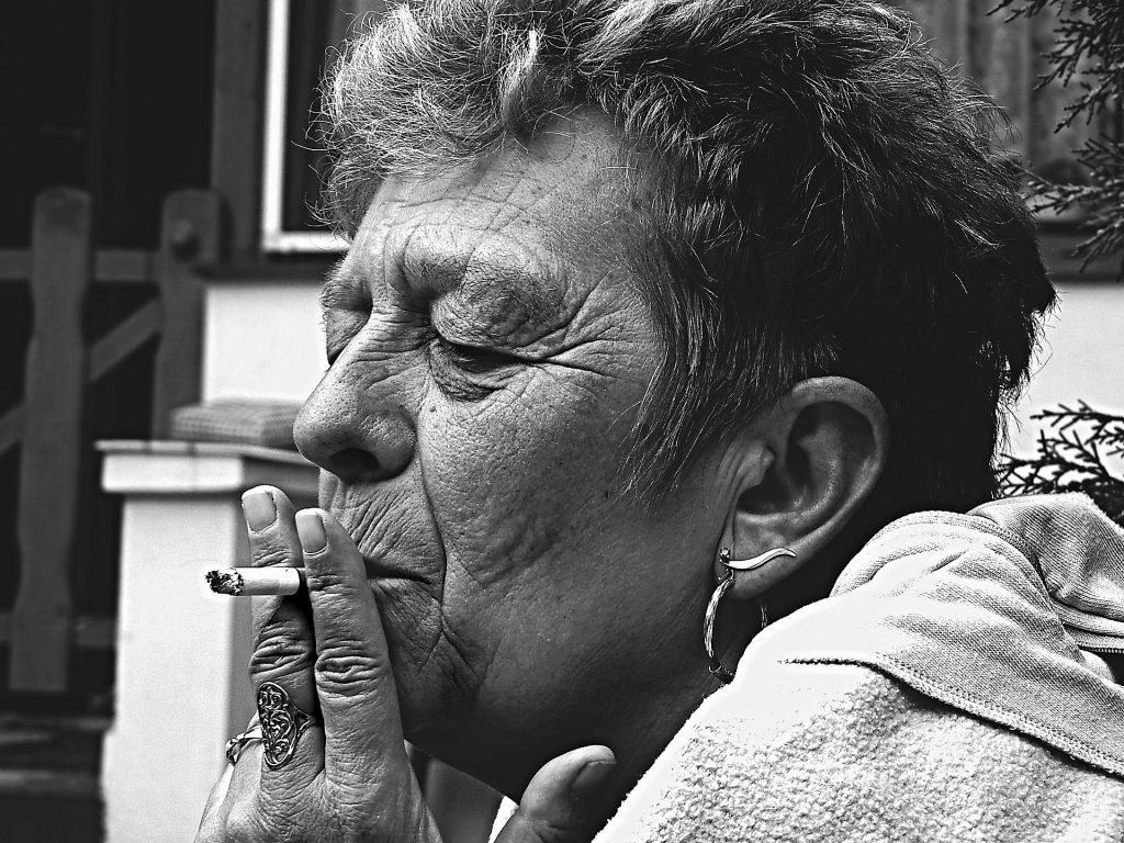 Imagem de uma senhora fumando. Ela está bem envelhecida. Tem cabelo curto, usa anel e brincos de prata.