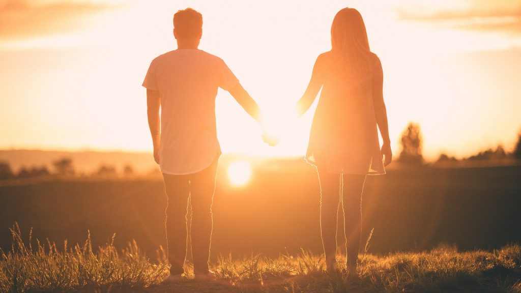 Imagem de um casal homem e mulher de mãos dadas olhando para o por do sol. Eles estão em uma montanha.
