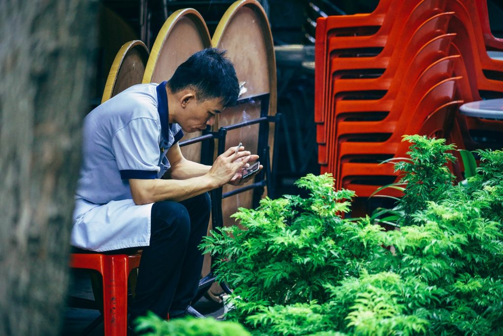 Imagem de um homem oriental sentado em uma cadeira vermelha. Ele está mexendo no celular e na outra mão segurando um cigarro aceso.
