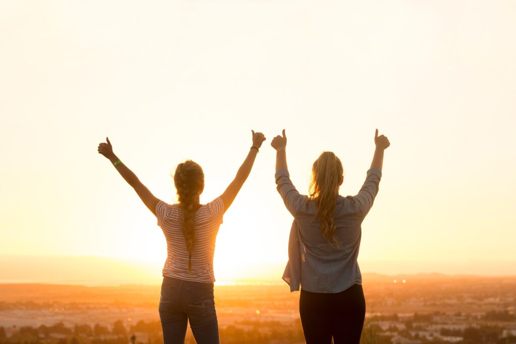 Imagem de duas mulheres de costas com os braços erguidos em sinal de positividade olhando para o por do sol.
