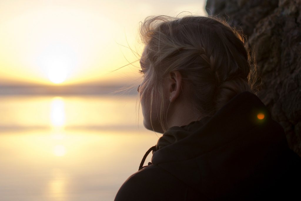 Imagem de uma garota usando tranças. Ela está encostada em um árvore olhando para o por do sol em frente ao mar.