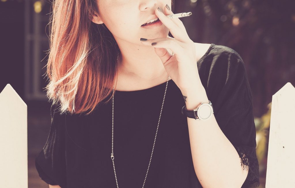 Imagem de uma jovem mulher de cabelos curtos e claros. Ela está fumando um cigarro. Usa blusa de manga curta e relógio com pulseira na cor preto.
