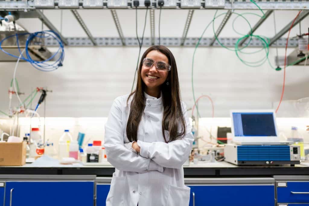 Mulher cientista com os braços cruzados sorrindo para foto dentro de uma laboratório. 