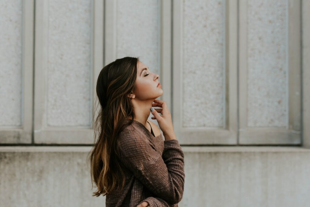 Mulher de cabelos lisos e suéter de lã vista de perfil, com os braços cruzados e a mão no pescoço, de olhos fechados.