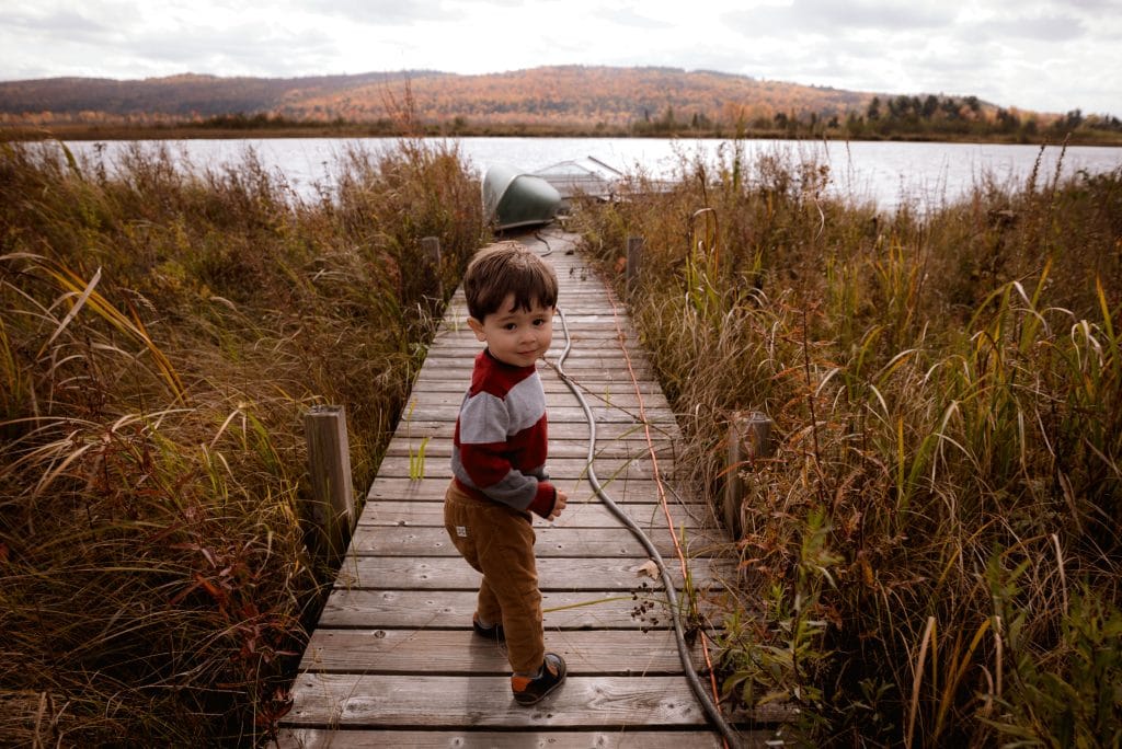 Bebê em pé em ponte com mato dos lados e lago ao fundo