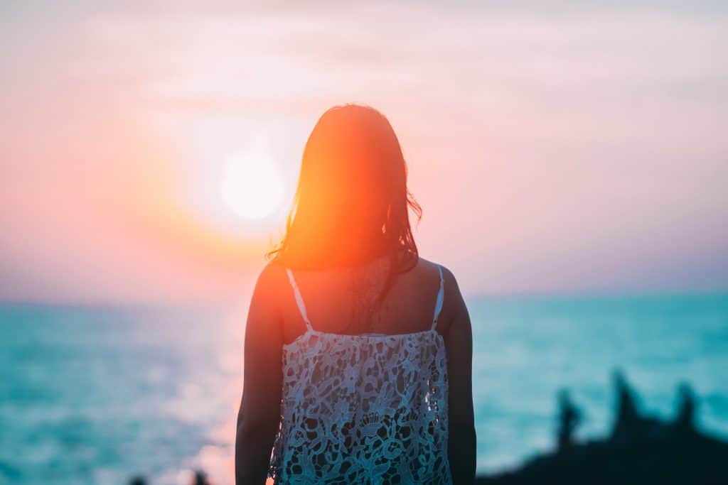 Mulher de costas com pôr-do-sol ao fundo refletindo e mar
