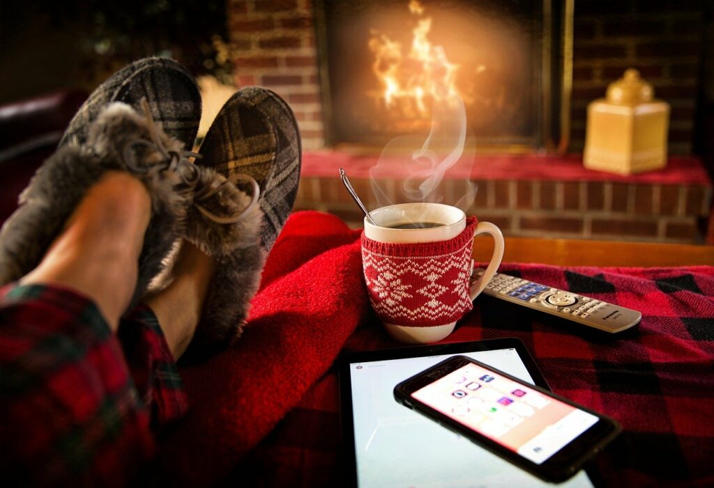 Pés com pantufas repousando em cima de uma mesa, com uma xícara quente ao lado, um tablet e um celular. Ao fundo, uma lareira acesa.