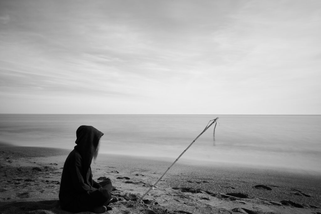 Imagem de uma mulher vestindo uma roupa preta. Ela está com um capuz preto sobre a cabeça e sentada na areia olhando para o mar. Ela está triste. A imagem é preto e branco.
