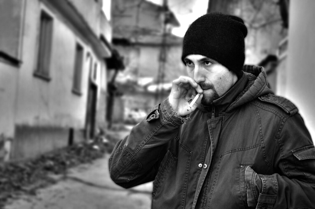 Imagem em preto e branco de um homem com barba cerrada. Ele está agasalhado com uma jaqueta de frio e uma touca de lã preta. Ele está em um beco de rua e fuma um cigarro.
