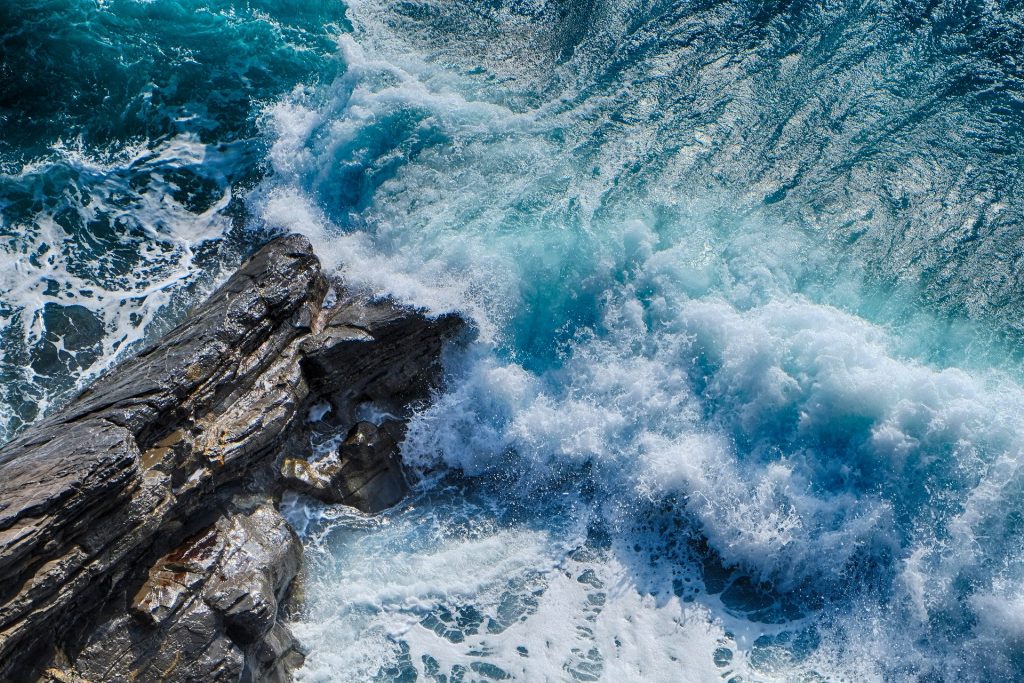 Imagem de uma praia azul. As ondas estão quebrando em uma pedra rochosa.