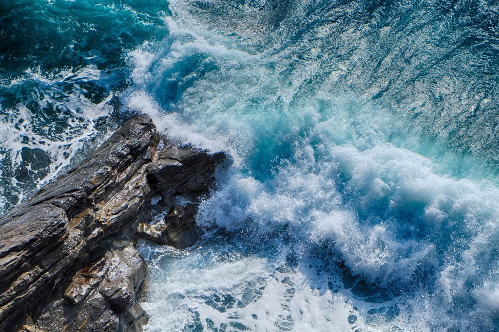 Imagem de uma praia azul. As ondas estão quebrando em uma pedra rochosa.
