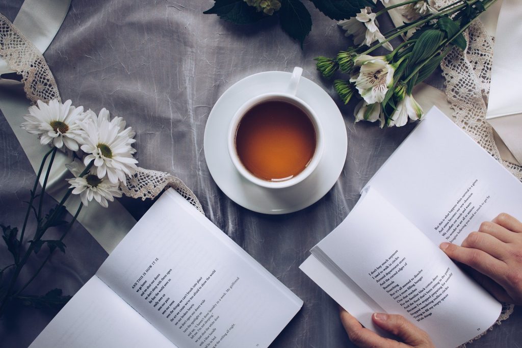 Imagem de uma xícara de porcelana branca cheia de chá. Ela está sobre uma mesa forrada com uma toalha cinza decorada com margaridas. Temos ainda dois livros abertos, sendo um deles manuseado por mãos de uma mulher.
