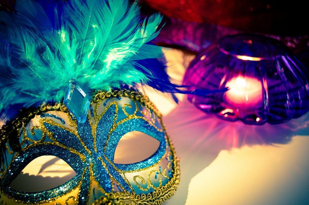 Máscara carnavalesca com detalhes em purpurina azul e amarela, e penas verdes, em cima de uma mesa ao lado de um vaso lilás com uma vela acesa dentro.