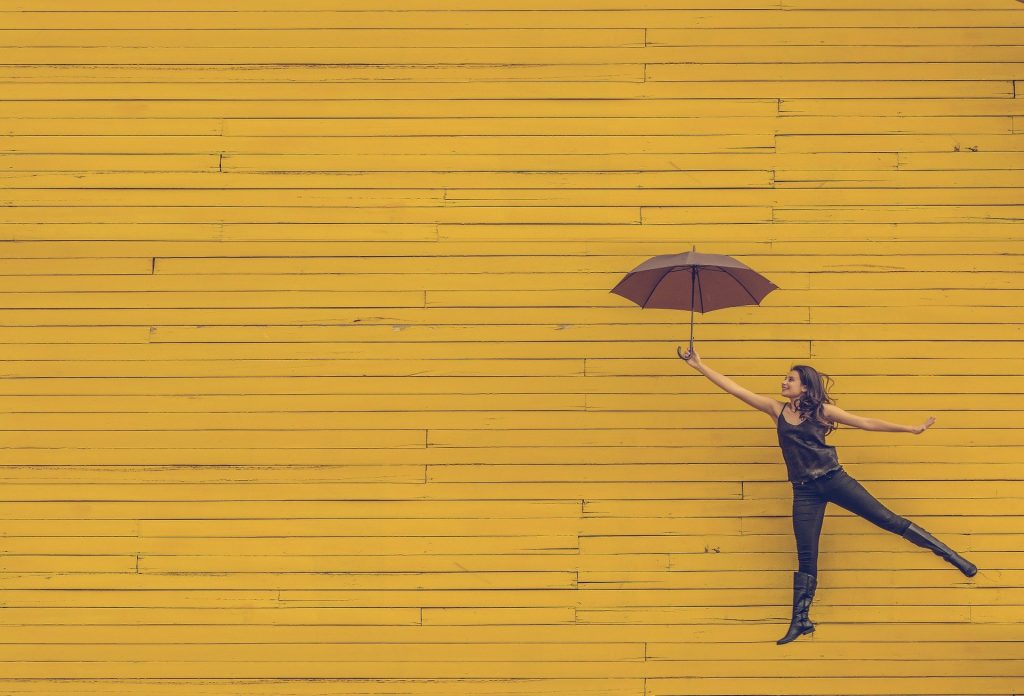 Imagem de uma mulher feliz vestindo uma roupa preta. Ela segura um guarda-chuva marrom. Ao fundo uma parede amarela.