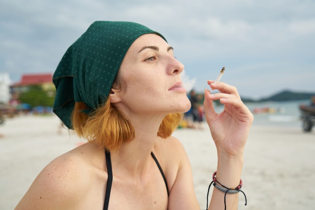 Imagem de uma mulher loira de cabelos curtos. Ela usa um lenço verde na cabeça, um biquini preto. Ela está na praia e fuma um cigarro.
