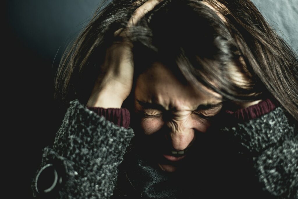 Mulher franzindo todo seu rosto, com os olhos fechados e a boca aberta como se estivesse gritando. Sua cabeça está baixa, e suas mãos estão em sua cabeça.