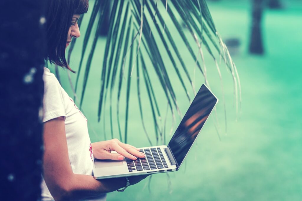 Mulher segurando notebook com a mão direita e digitando com a mão esquerda, embaixo de uma palmeira.