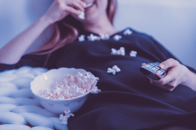 Garota deitada na cama comendo pipoca segurando controle da TV e sorrindo