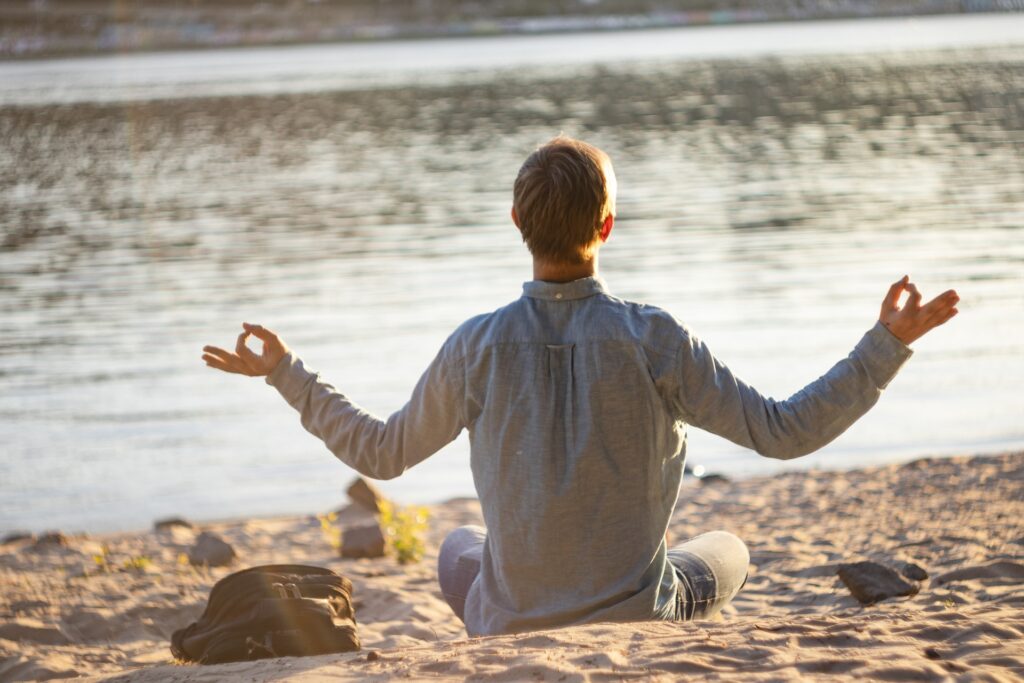 Imagem de um homem em posição de meditação. Ele está sentado sobre a areia de uma praia olhando para o mar.
