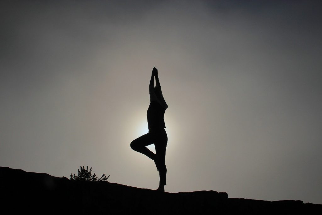 Silhueta de uma mulher em posição de yoga. Ela está em pé, com as dois braços erguidos e ao fundo a imagem do por do sol.