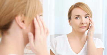 Mulher de meia idade olhando para o seu reflexo no espelho enquanto toca seu rosto, com expressão séria.