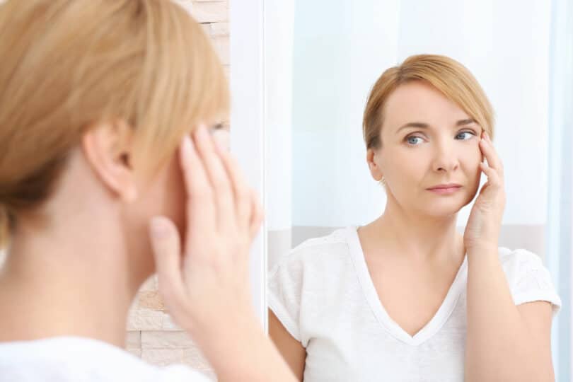 Mulher de meia idade olhando para o seu reflexo no espelho enquanto toca seu rosto, com expressão séria.