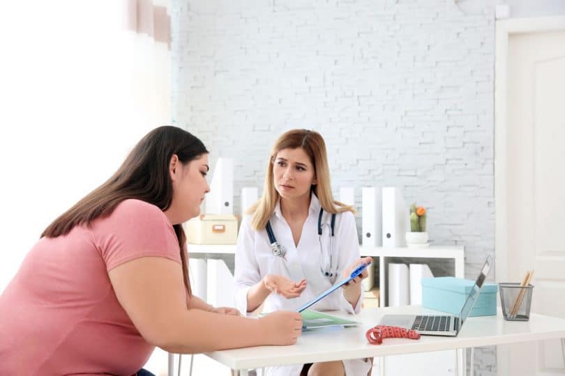 Mulher obesa em consulta com médica, que lhe apresenta informações em uma prancheta.